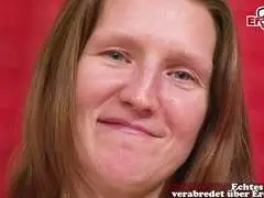 Reife brünette Milf sitzt brav mit verschränkten Händen auf rotem Sofa