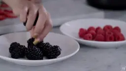 Hand legt eine Brombeere auf schwarze Beeren in weißer Schale, daneben Himbeeren auf Marmor.