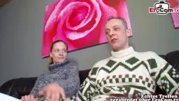 Schlanke blonde Ehefrau sitzt lächelnd zwischen zwei Männern auf schwarzem Ledersofa im Wohnzimmer