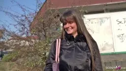 Junges brünettes Mädchen mit langen Haaren und schwarzer Jacke lächelt scheu vor Berliner Plattenbau