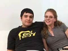 Nerdgirl mit Brille und ihr Freund sitzen eng umschlungen auf weißer Couch, beide lächeln in die Kamera.